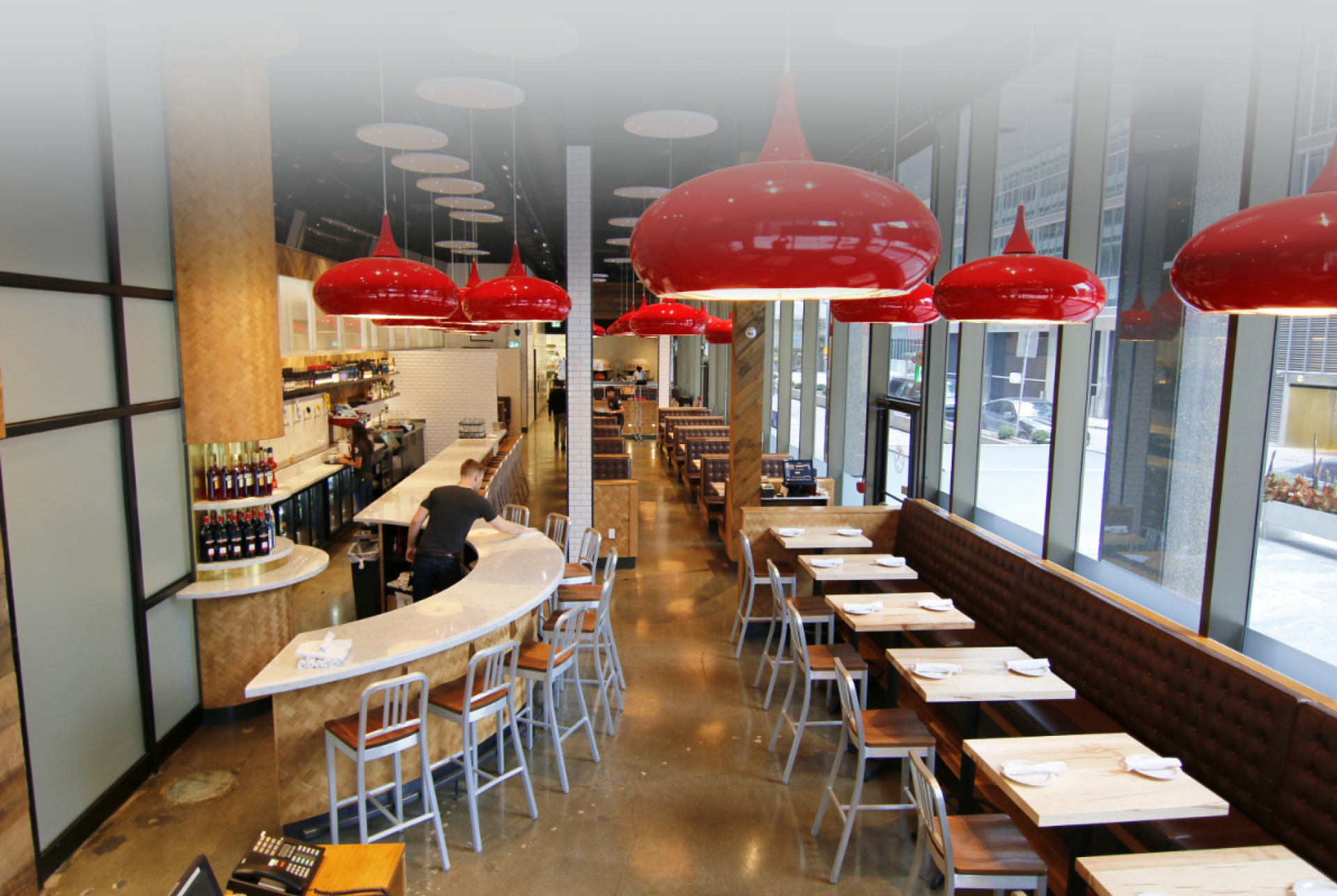 A modern restaurant interior with a curved bar, wood tables, and bench seating along large floor‑to‑ceiling windows. Red pendant lights hang from a dark ceiling, adding contrast to the bright, contemporary dining space.